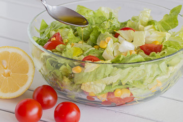 A glass bowl with green salad, cherry tomatoes and corn. A spoon with olive oil.