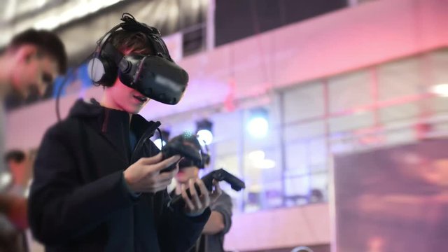 The guy plays in the VR headset virtual reality at a meeting events of computer game fans. In the background there are a lot of people in the blur, bright and lights from concert.