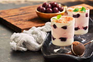 Layered dessert in glasses with yoghurt and canned cherries, sprinkled with cocoa powder and crumpled with mint leaves.