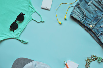 Top view of summer clothing and accessories smartphone, top, necklace and black sunglasses on light blue background with copy space for text. Summer is coming concept.