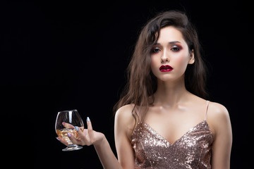 Beautiful glamorous brunette girl wearing a sparkling dress sensually holds in her hand a glass of whiskey with ice.