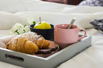 Breakfast in bed. A cup of coffee, croissants on a tray, an apple and a flowers.