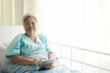 Elderly patient using tablet in bed. Elderly chinese woman in hospital bed using tablet to connect with her relatives. Connected world concept.