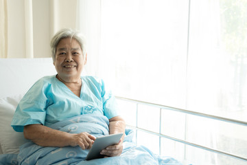 Elderly patient using tablet in bed. Elderly chinese woman in hospital bed using tablet to connect with her relatives. Connected world concept.