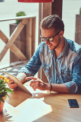 Hipster guy with a stylish haircut and beard sits at a table in a roadside cafe, using a tablet computer.