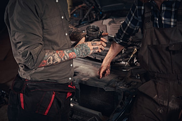 Obraz premium Two mechanic examine a detail of the engine while standing near a broken car in a garage.