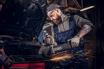 Two bearded auto mechanic in a uniform and safety glasses working with an angle grinder while...