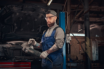 Fototapeta na wymiar Brutal auto mechanic in a uniform and safety glasses working with an angle grinder while standing against a broken car in repair garage.