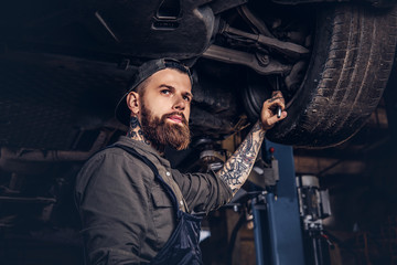 Bearded auto mechanic in a uniform repair the car's suspension with a wrench while standing under...