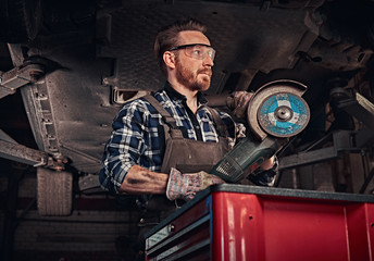 Fototapeta na wymiar Mechanic in a uniform and safety glasses holds an angle grinder while standing under lifting car in a repair garage.