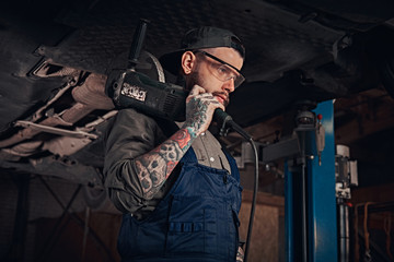 Mechanic in a uniform and safety glasses holds an angle grinder while standing under lifting car in a repair garage.