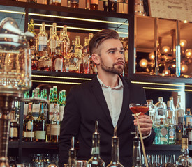 Handsome stylish male holds a glass of exclusive alcohol standing at bar counter background.