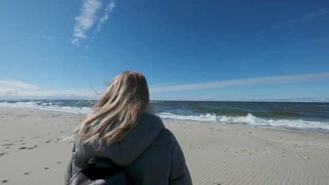 Lovely blonde girl steps on a sandy beach on a background of blue sea waves, blue sky. Slow motion