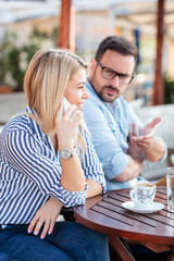 Happy young woman talking on the phone, ignoring her boyfriend in background. Relaxing and drinking coffee in a cafe. Selective focus
