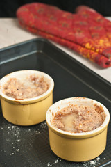 Top view of two freshly baked, yellow ramekins , filled with apple, raisin, sour cream crumble cooling on a backing sheet on a wood table top with one oven glove