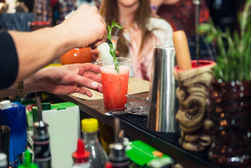 Expert pro barman hands is making cocktail and decorating drink with fresh rosmarine on the color background with people. Drinking cocktails, nightlife and party event concept. Sective focus.