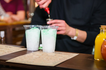Expert pro barman is making cocktail and pouring drink into a plastic glass on the color background with people. Drinking cocktails, nightlife and party event concept. Sective focus, space for text.