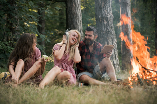 Friends relax at bonfire flame with sparks in vintage style. People camping at fire in forest. Women and bearded man at campfire. Eating food, reading book and entertainment. Summer vacation concept