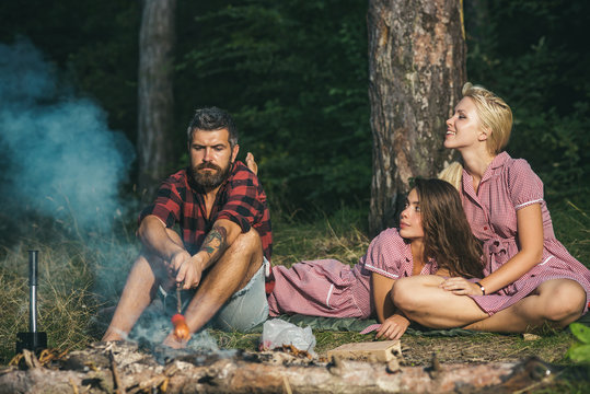 Friends camping in forest. Two playful girls in vintage dresses looking at lumberjack. Guy with serious look sitting by campfire and flying sausages
