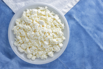 Cottage cheese on a white blue fabric background, soft cheese on a white plate, fresh homemade cheese on a white blue napkin. Dairy product in vintage style, healthy lifestyle. Copy space