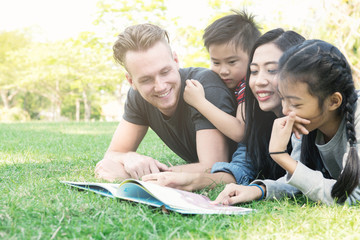 Family reading a book together in park. Mixed race family laying down together reading book. White man, chinese woman, chinese boy and girl. Low angle shot.