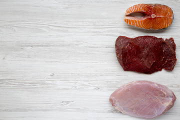 Uncooked raw salmon steak, beef meat and turkey breast on white wooden background, top view. Flat lay. Copy space.