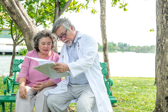 Doctor With Patient In Park. Senior Chinese Male Doctor Sitting Together With Chinese Female Patient. Talking And Examine Her.