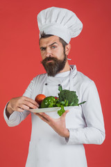 Chef with serious face in white uniform holds vegetables on plate. Cooking and vegetarian diet concept. Green grocery concept. Male chef in cook uniform holds vegetable plate in hand.