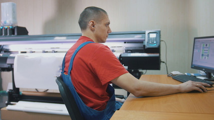 Male worker in typography with computer in front of printing press