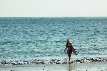 Fototapeta premium back view of female surfer with surfboard walking on beach into the ocean