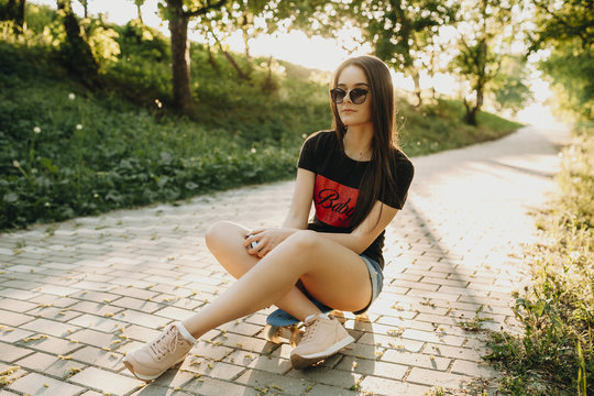 Full length portrait of a lovely young girl sitting on a small skateboard wearing sunglasses and looking into camera .