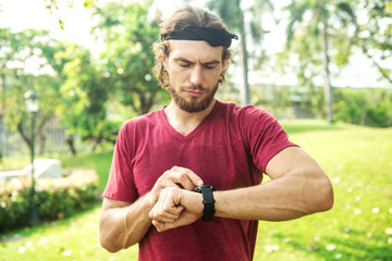 Naklejka na ściany i meble Fitness man using a smart watch in park. White male pressing button on his smart sport watch to keep track of his fitness activties.