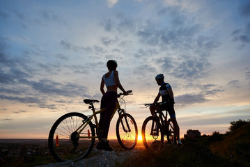 Fototapeta premium Two young sports people on mountain bikes stand on rock on top of hill looking at the sun at sunset. Girl and guy dressed in helmets and sportswear stand facing each other under evening sky