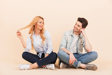 Image of caucasian couple man and woman 20s in denim clothing smiling while sitting on the floor with legs crossed, isolated over beige background