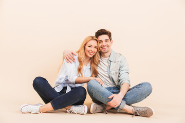 Image of nice and cute couple in denim clothing smiling and hugging together while sitting on the floor with legs crossed, isolated over beige background