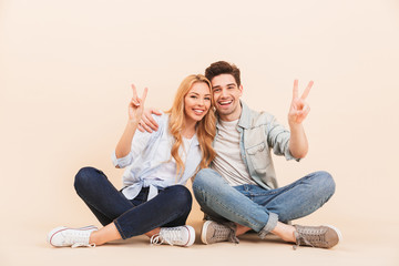 Image of pleased man and woman hugging together while sitting on the floor with legs crossed and showing peace symbols, isolated over beige background