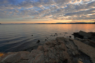 Krajobraz morskiego wybrzeża podczas zachodu słońca, skały o poszarpanej strukturze na pierwszym planie, lekko pomarszczona tafla wody, niebo z niskimi chmurami, horyzont rozświetlony promieniami