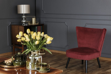 Red armchair in dark grey living room interior with lamp and yellow flowers on table