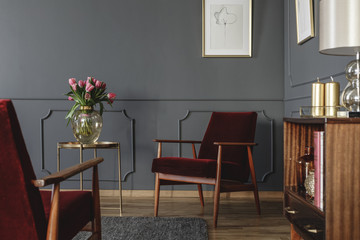 Pink flowers on gold table next to red wooden armchair in dark living room interior