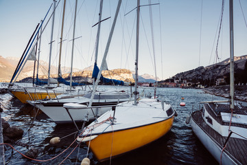 Boats moored in the harbor in Italy on the surface of the mountain lake Lago di Garda during the summer sunrise with town and clear sky in the background