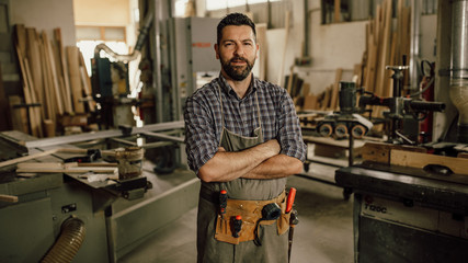 Portrait of a smiling young woodworker standing with his arms crossed by a bench saw in his workshop full of carpentry equipment - obrazy, fototapety, plakaty