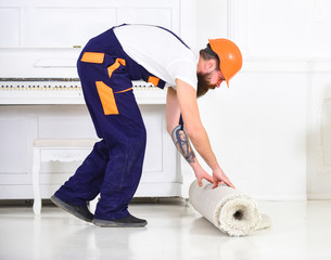 Relocating concept. Man with beard, worker in overalls and helmet rolling carpet, white background. Loader wrappes carpet into roll. Courier delivers furniture in case of move out, relocation.