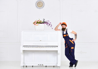Man with beard, worker in overalls and helmet lean on piano, white background. Loader moves piano...