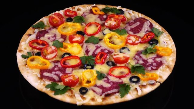 Whole handmade tasty pizza with salami and vegetables just from oven rotating against black background. 4K. Traditional tasty food.

