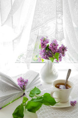 white cup with tea and a bouquet of lilac in a white vase on a summer morning breakfast at the window with a white lacy curtain in a rustic style with a copy of the space