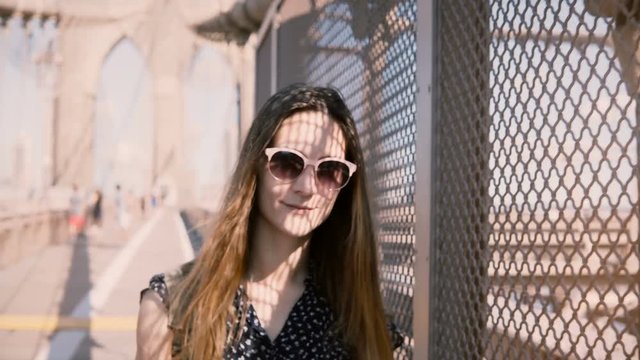 Beautiful female tourist in sunglasses smiling, leaning to shadow net fence at Brooklyn Bridge, walking to camera 4K.