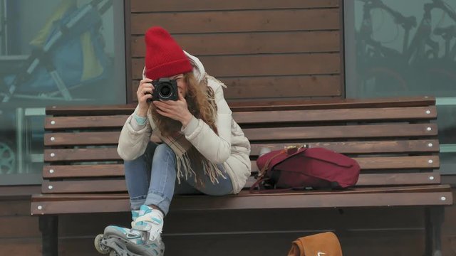 Young beautiful woman in a red hat wearing sporty warm clothes and rollers, sitting on a wooden bench and taking pictures on a vintage camera