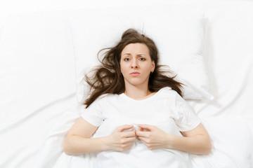 Top view of tired stressed crying young woman lying in bed with white sheet, pillow, blanket. Pensive frustrated sad upset female spending time in room. Rest, relax, bad mood concept. Copy space.