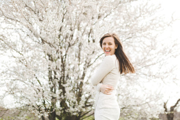 Young happy laughing beautiful woman in light casual clothes holding hands folded standing in city garden or park on blooming tree background. Spring nature, flowers. Lifestyle, leisure concept.
