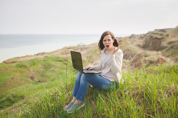 Fototapeta premium Young irritated dissatisfied business woman or student in casual clothes sitting on grass using laptop in field spreading hands working outdoors on green background. Mobile Office. Lifestyle concept.